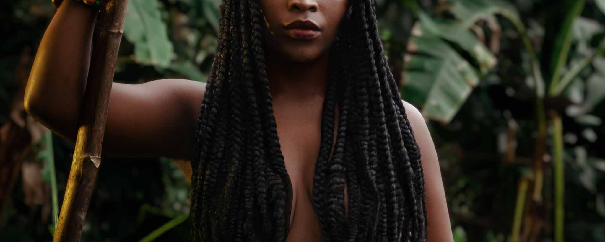 'Long dreadlocks cover the bare-chest of native female holding a staff while standing in front of foliage.'