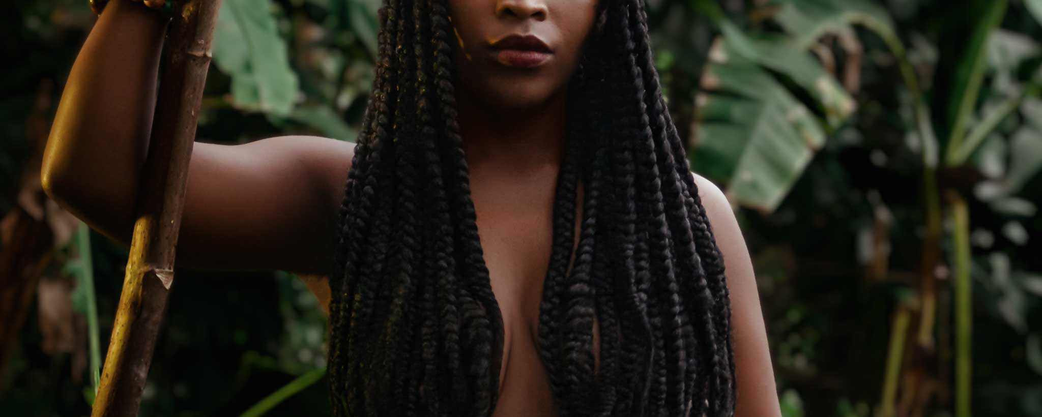 'Long dreadlocks cover the bare-chest of native female holding a staff while standing in front of foliage.'