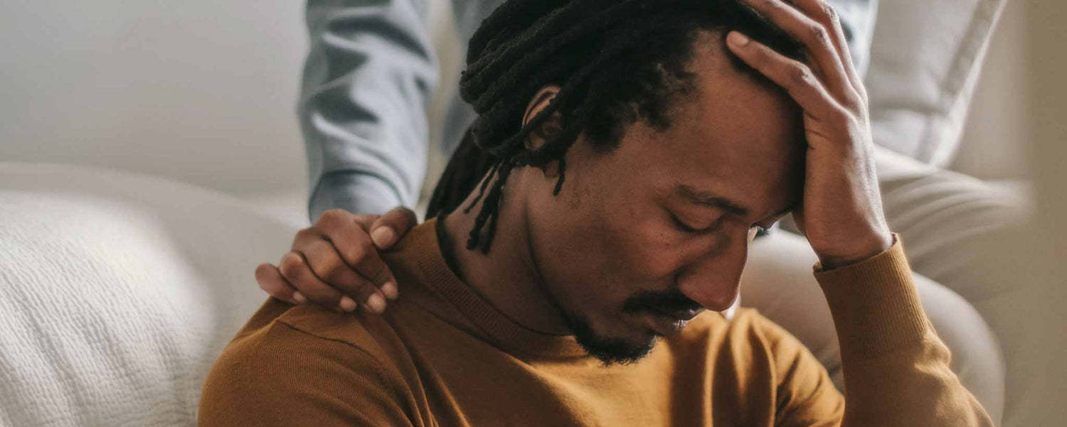 A man in despair holds his head while a female offers a comforting hand on his shoulder.