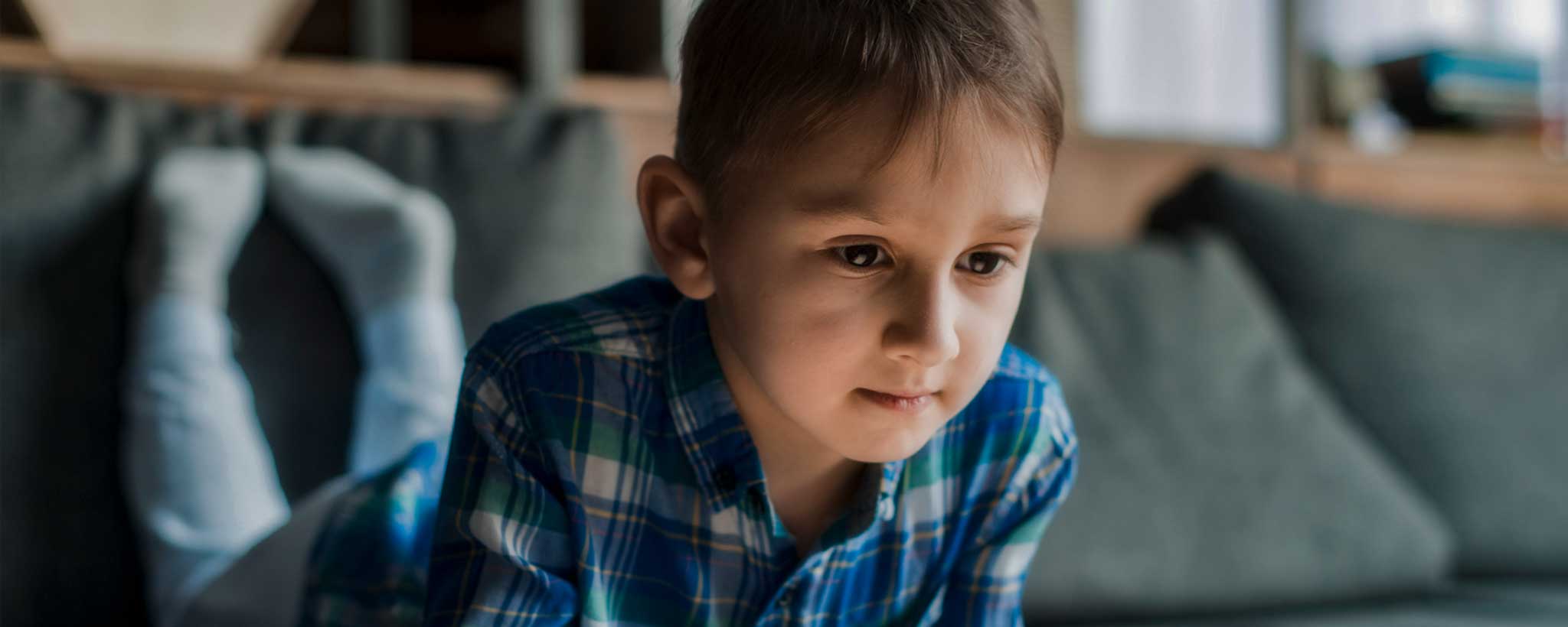 'A boy lies prone on a sofa while staring at a digital tablet.'