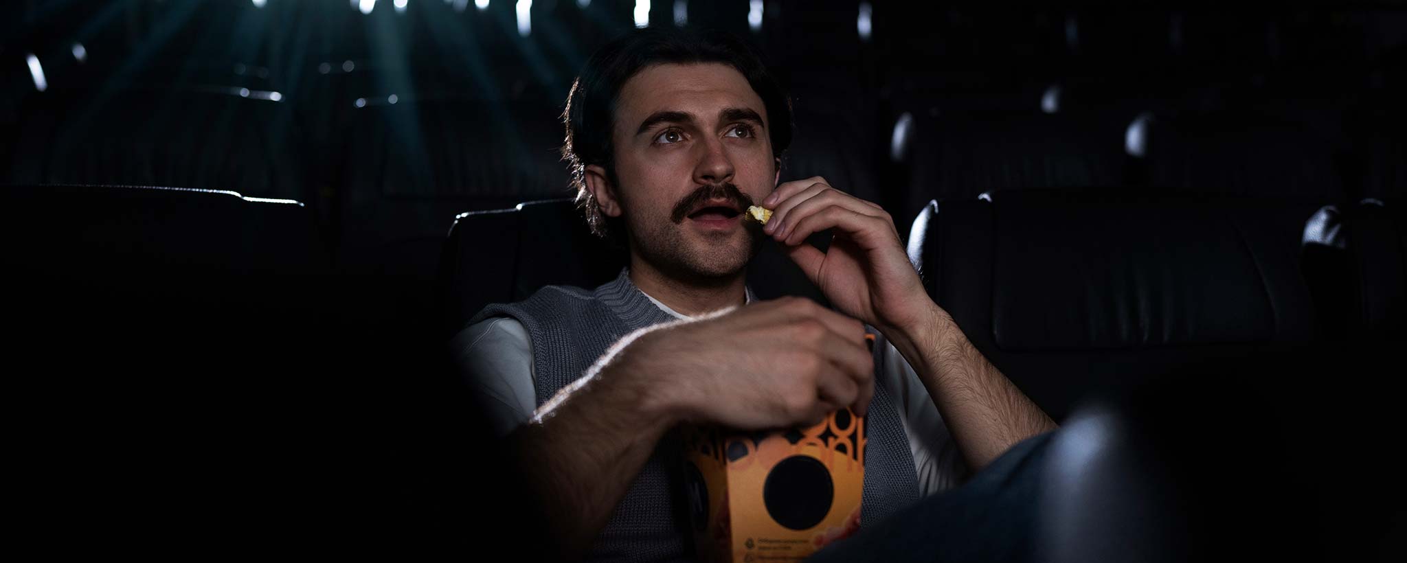 'A man is alone, eating popcorn in a movie theater with dim lighting.'