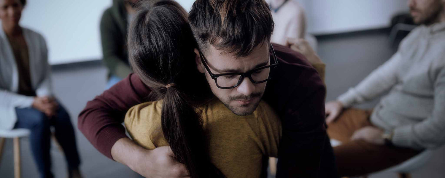 A man and woman comfort each other with a warm embrace while others are seated nearby.