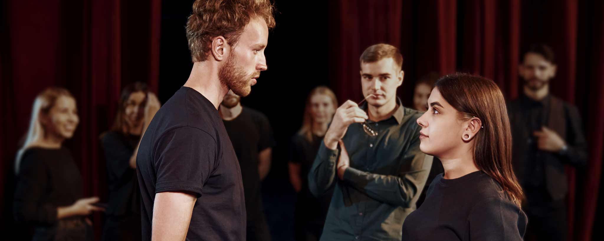 'A group of young men and women in black clothing standing on a stage in front of red curtains, with two central people facing each other in a rehearsal or performance setting.'