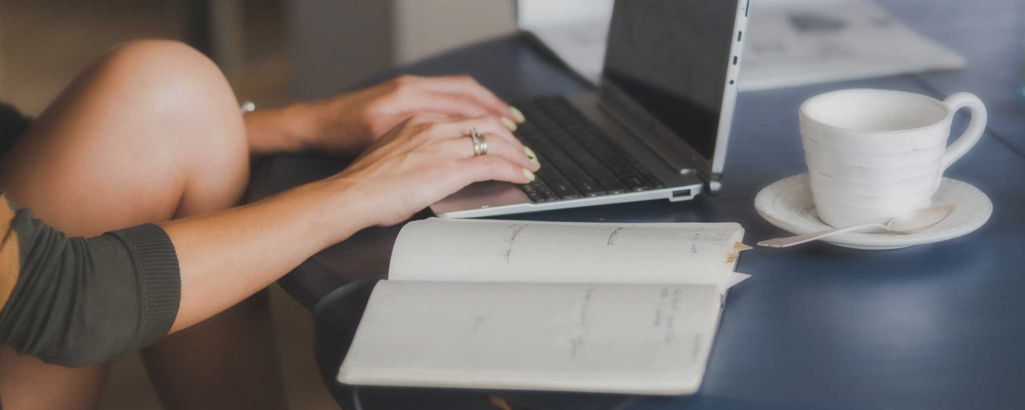 'Woman typing on a a laptop computer with an open book.'