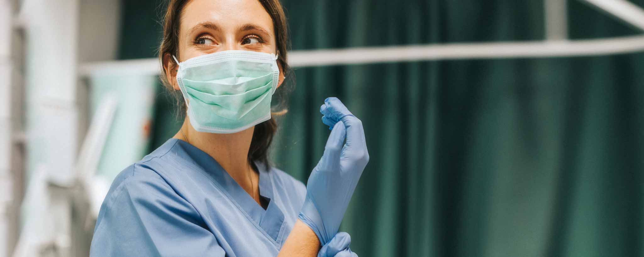 'A female medical professional wearing medical scrubs, a mask, and gloves in front of a blurred background.'