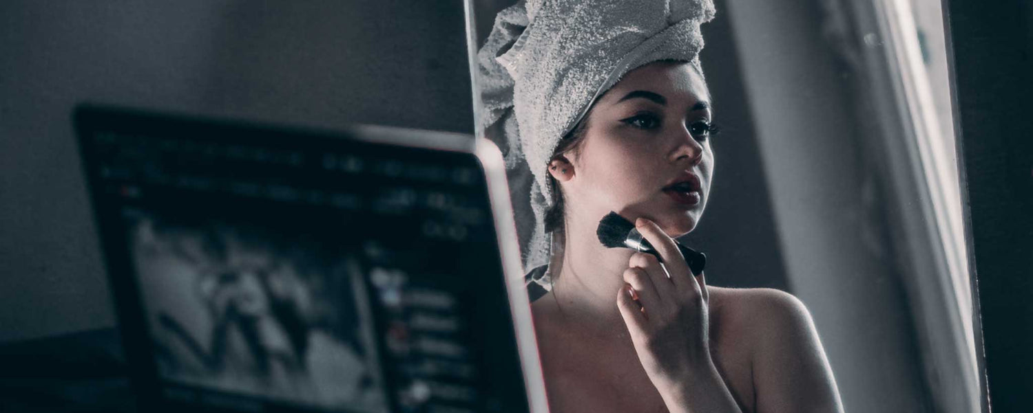 A lady applies makeup in a mirror with her laptop in the foreground.