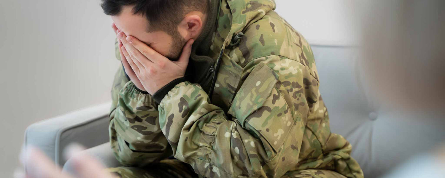 A man wearing a military uniform is sitting on a couch with his head in his hands, experiencing stress.