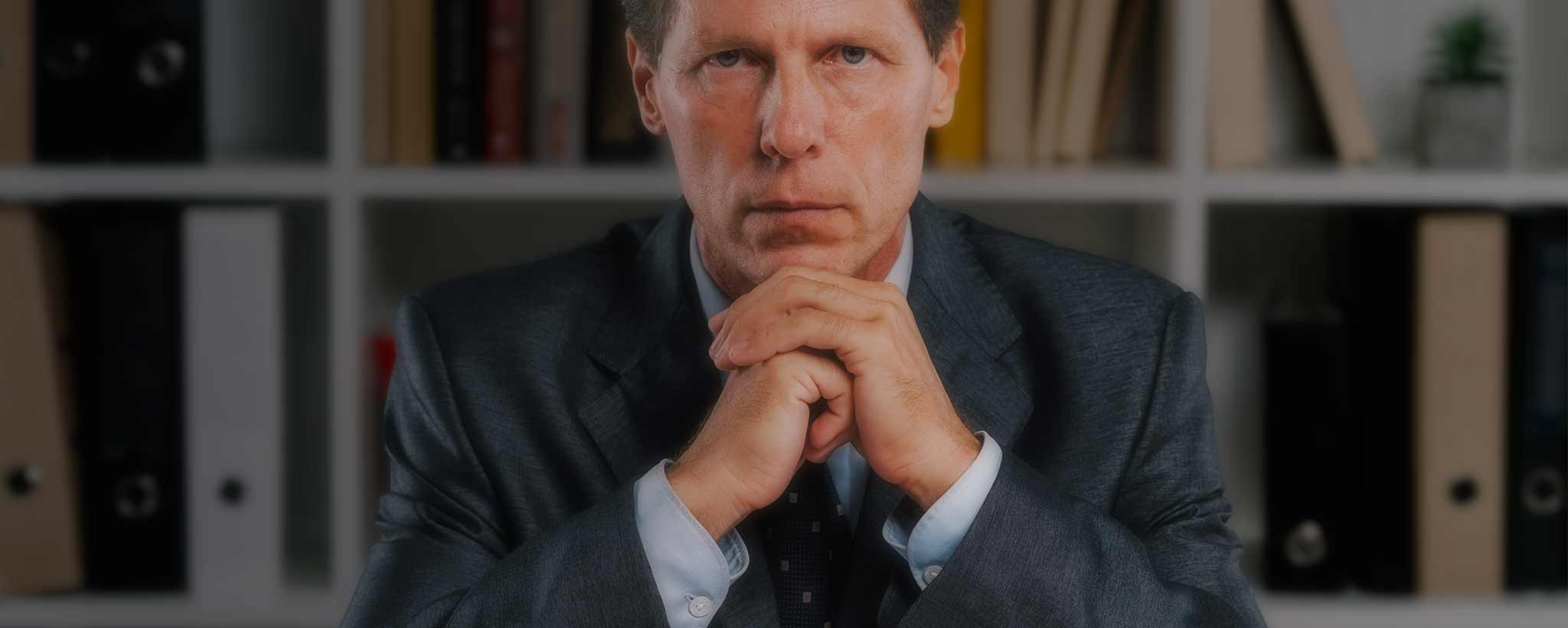 'A serious male attorney sits behind his desk with his hands clasped.'