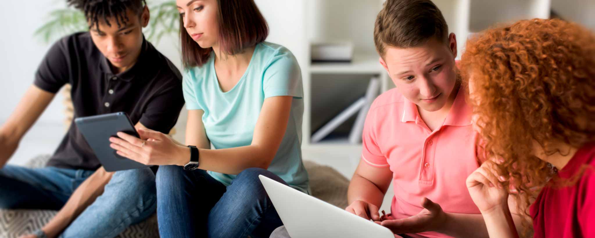 'Four students are sitting together, each using a different device in a casual setting.'