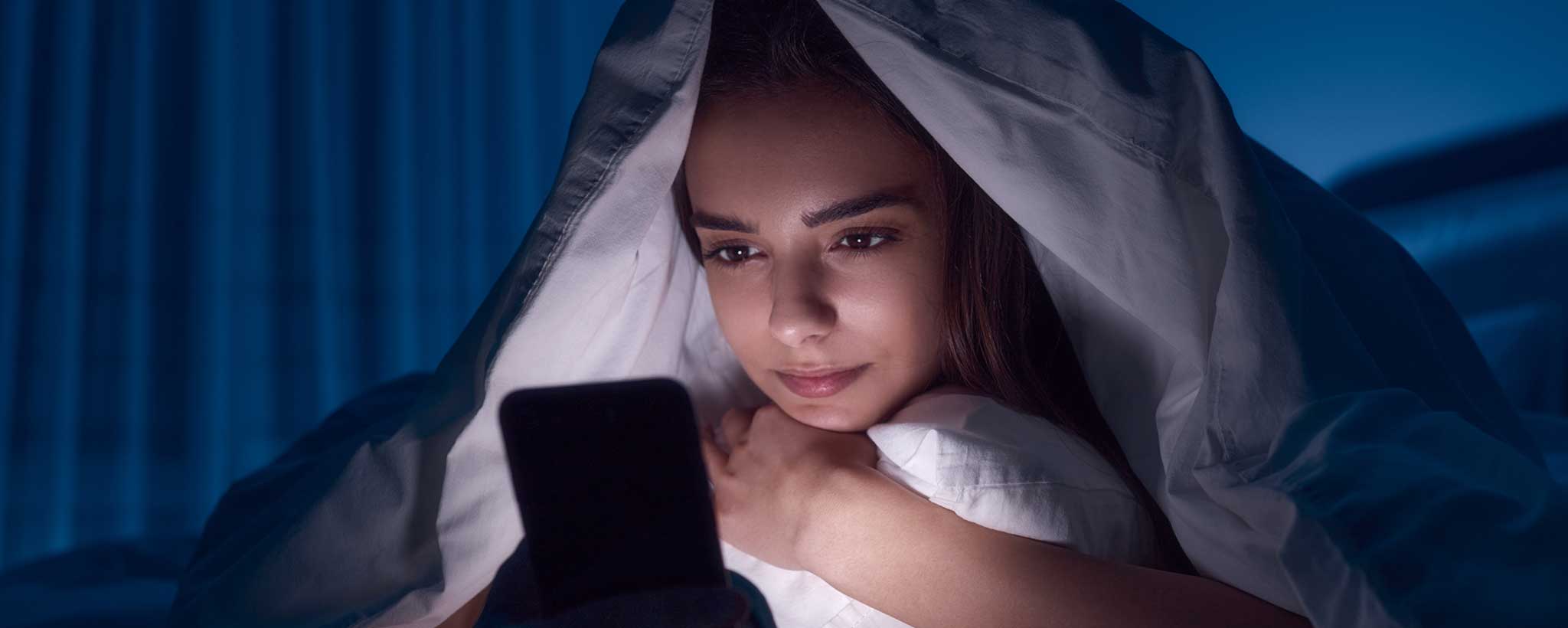 'A female looks at her phone beneath her bed sheets.'