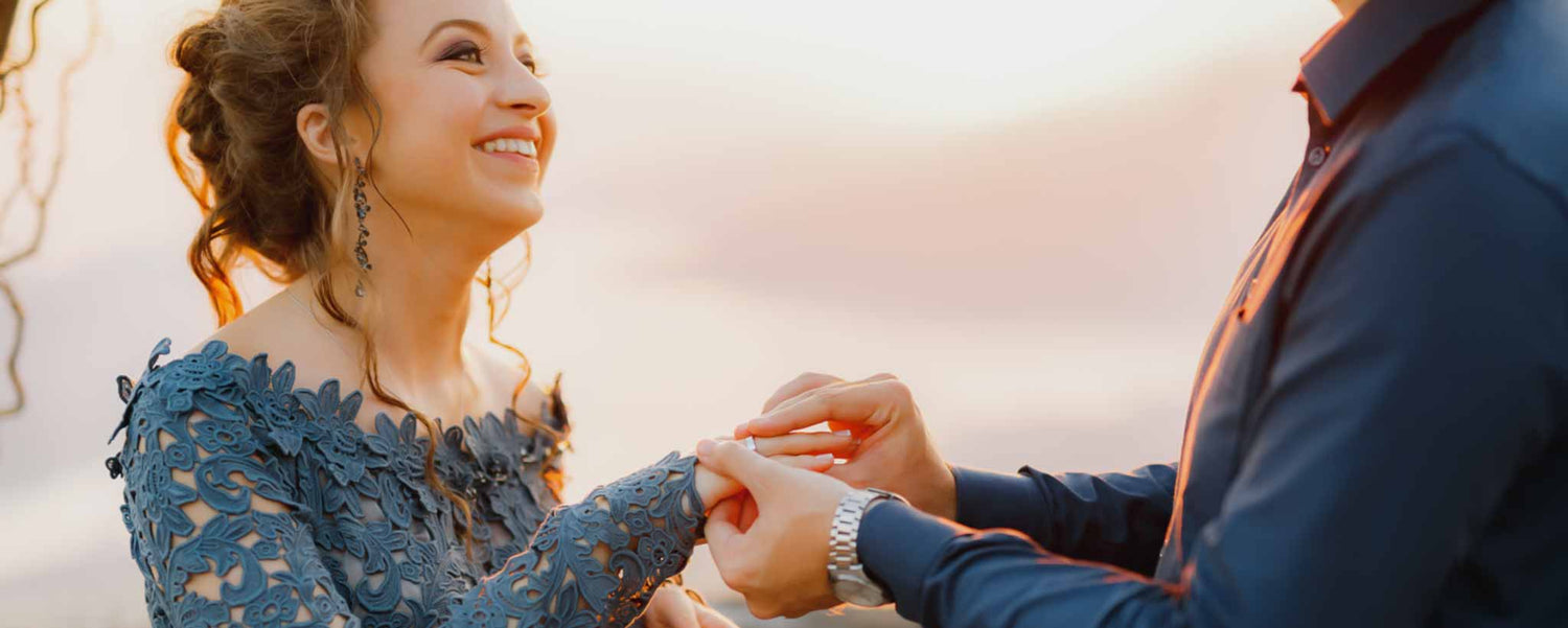 A happy lady in a blue dress receiving a ring from a man outdoors with a warm glow.