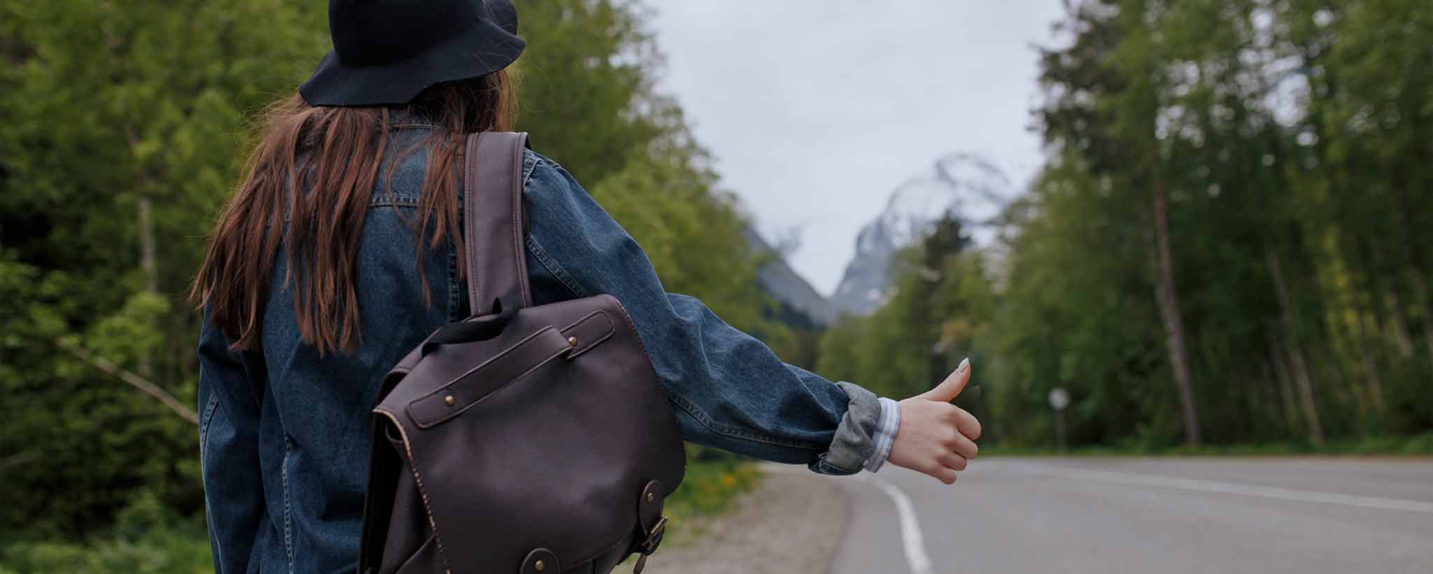 'Hitchhiking female with backpack stands near an empty road.'
