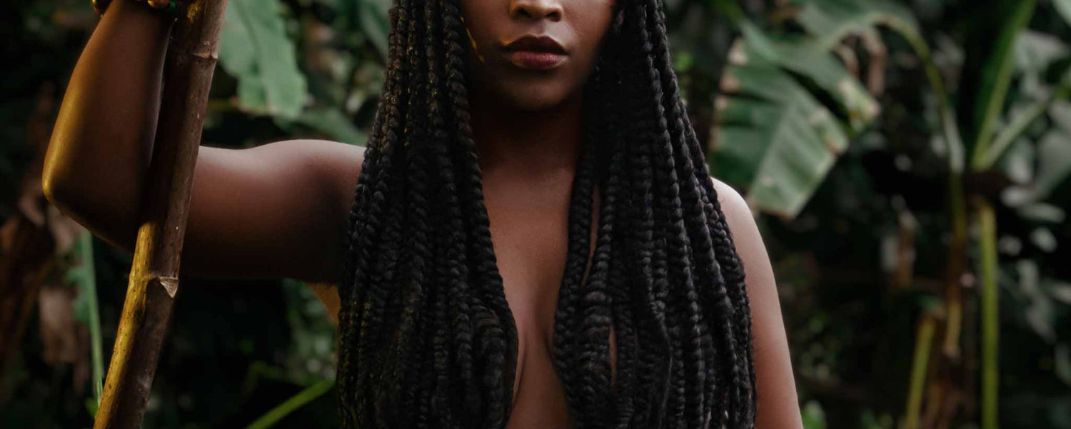 Long dreadlocks cover the bare-chest of native female holding a staff while standing in front of foliage.