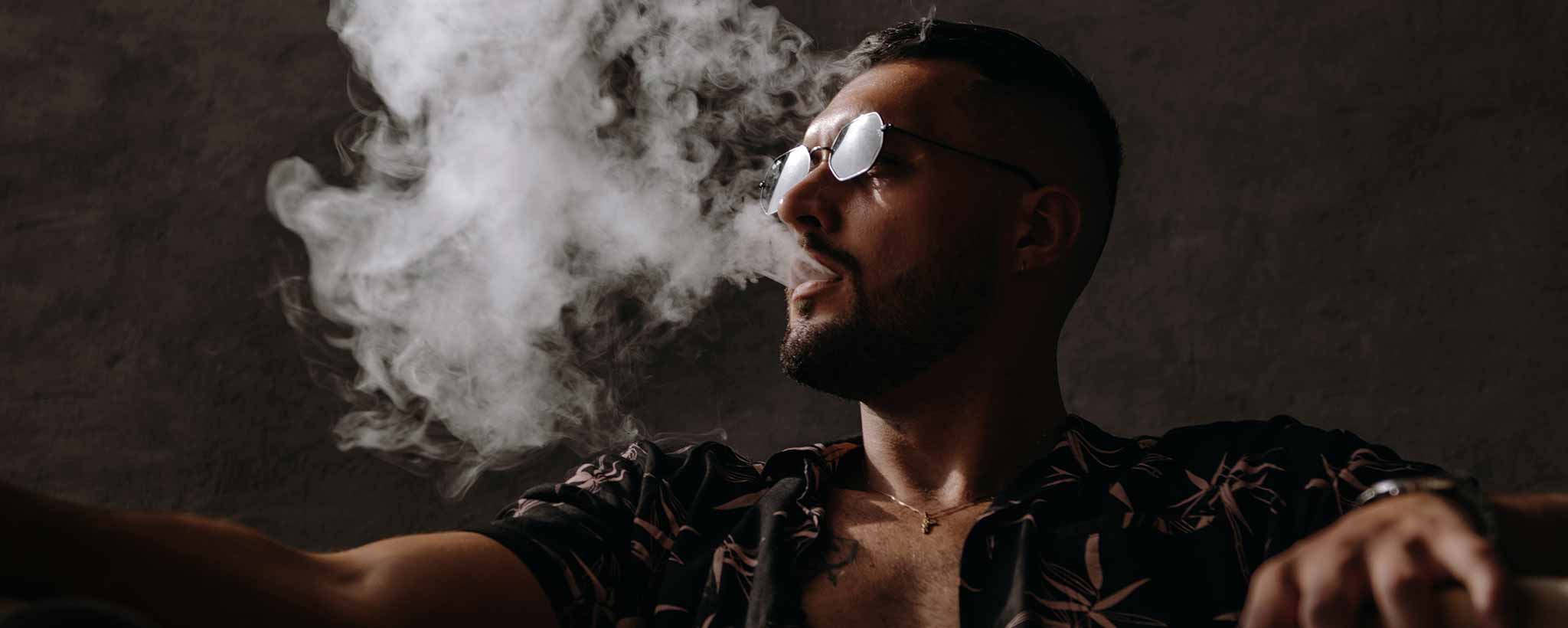A man with glasses pin a dark room blows white smoke.