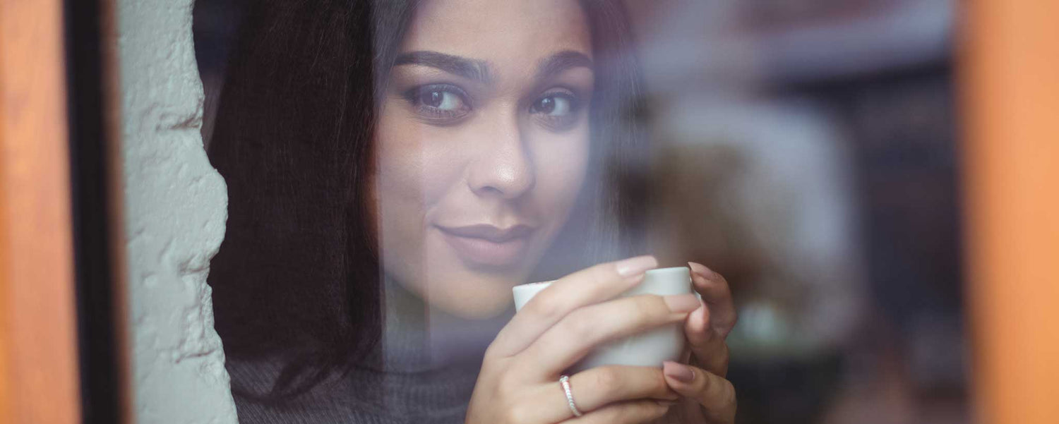 A lady holding a cup, looking through a window with a blurred background.