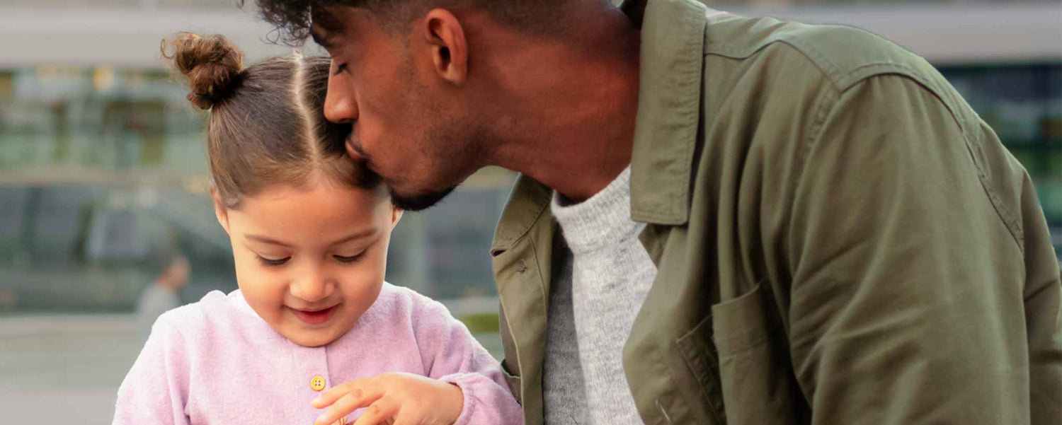 A black father kissing his white daughter outdoors.