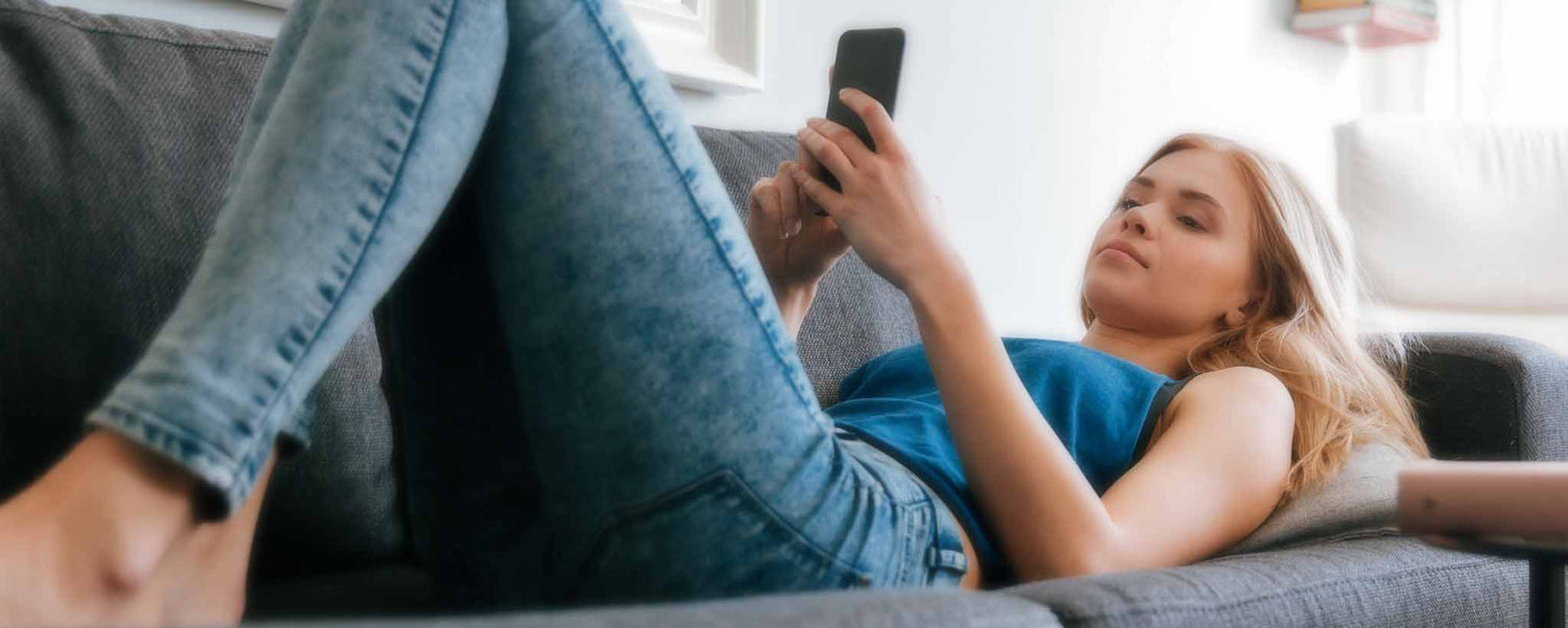 A female with a smartphone lies supine on her sofa, phone in hand.