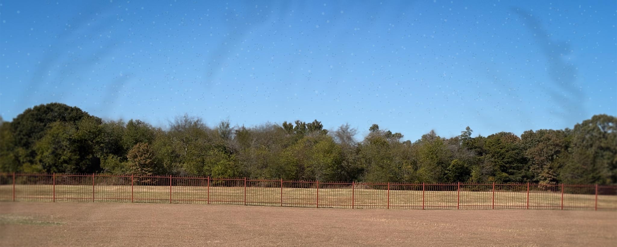 'Open field with a fence and trees under a clear blue sky with floaters.'