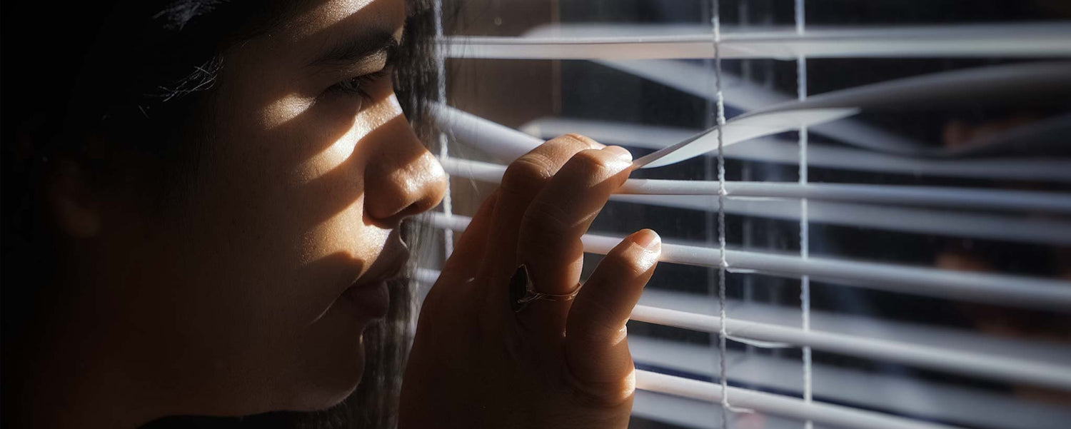 A person looking through window blinds in a dimly lit room.