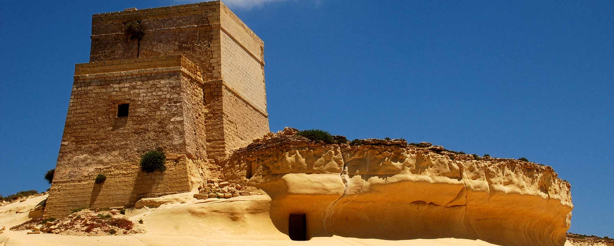 'A tall tan stone tower beside a large sandstone rock formation under a clear blue sky in Malta.'