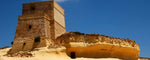 A tall tan stone tower beside a large sandstone rock formation under a clear blue sky in Malta.