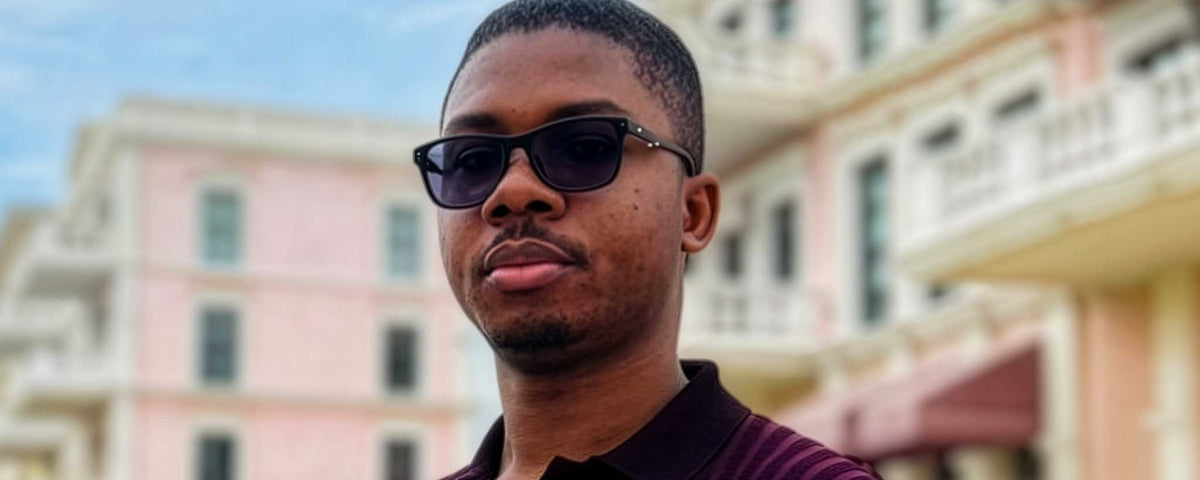 Portrait of Michael Nwokasu wearing black sunglasses and a dark maroon polo shirt, standing outdoors in Lagos with a blurred pink building in the background.