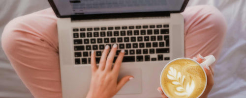 Woman sits with laptop on her lap and cappuccino in her hand