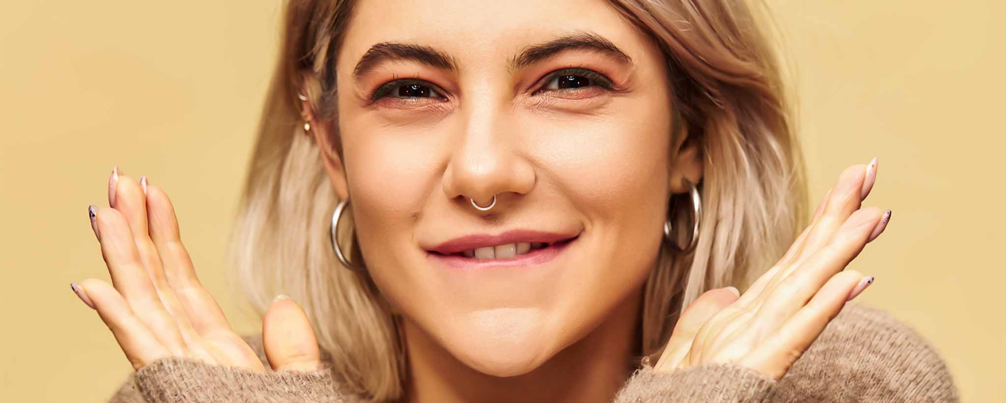 'A blonde female with a nose ring and earrings against a beige background.'