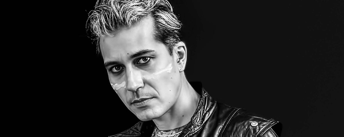 A black-and-white portrait of Reza Sixo Safai with styled hair, wearing a leather jacket against a dark background.
