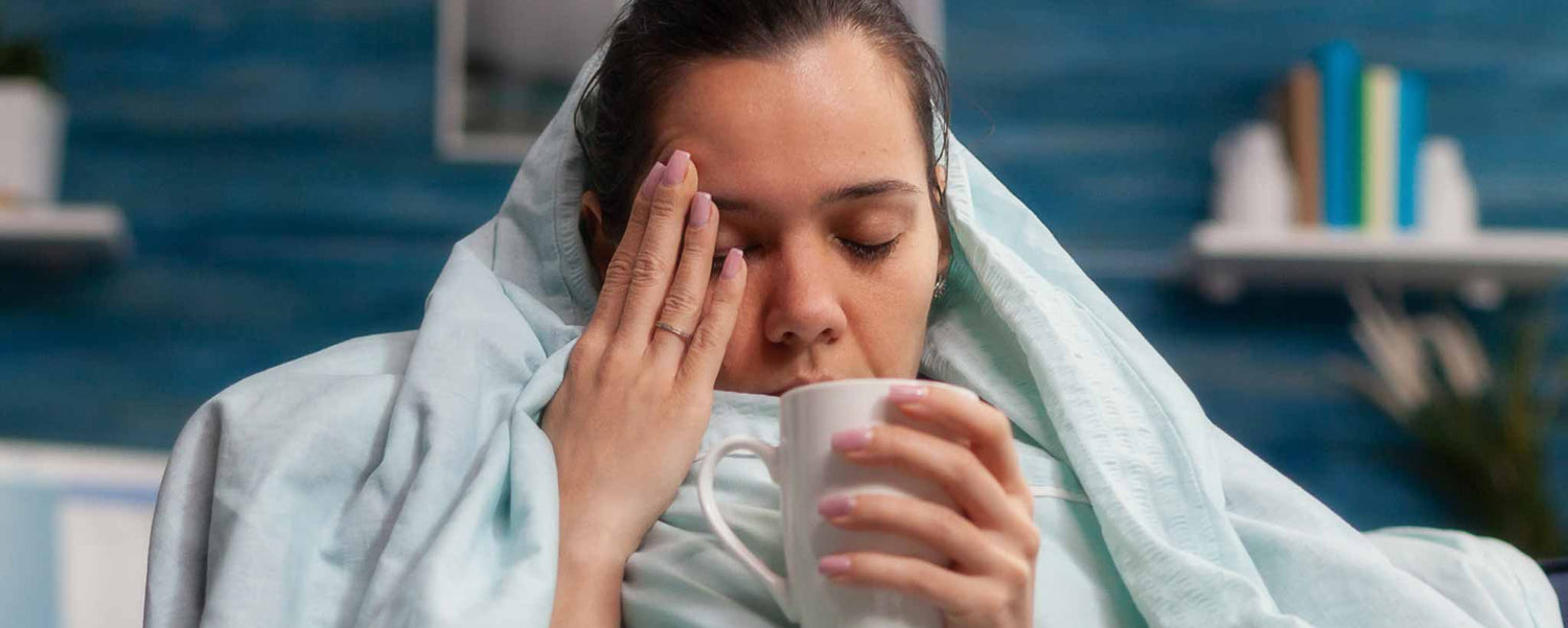 A lady in a blanket, holding a mug, and rubbing their face in a room with a blue wall.
