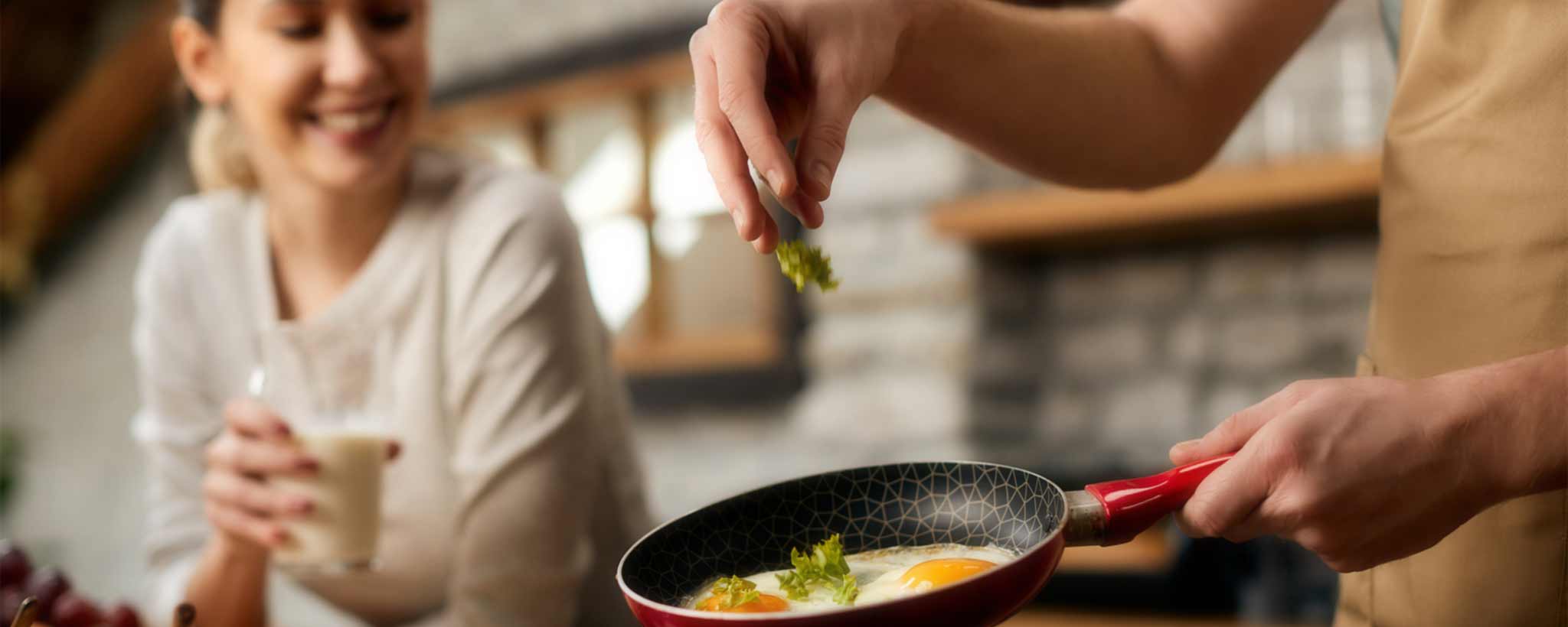 'A man fries eggs in a skillet while a woman looks on.'