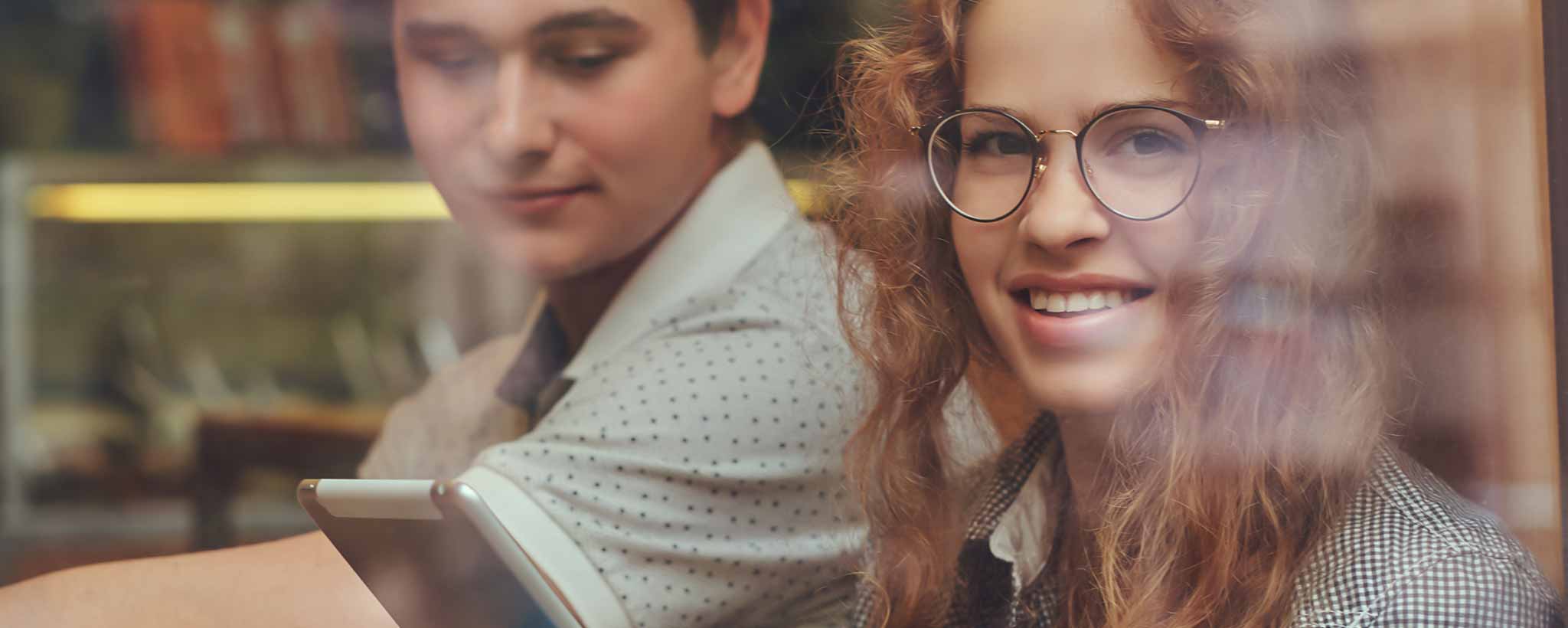 'Two people sitting together, one of whom is holding a digital tablet, wearing glasses, and smiling.'